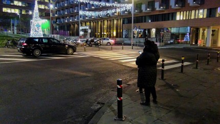 Person standing at the side of a street at night looking at their mobile device. Pedestrian crossing and a car in the background. Person standing at the side of a street at night looking at their mobile device. Pedestrian crossing and a car in the background.