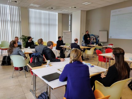 People sitting in groups of 4 in a well lit room at desks with their laptops and notebooks open, listening to a man giving a presentation People sitting in groups of 4 in a well lit room at desks with their laptops and notebooks open, listening to a man giving a presentation
