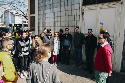 People standing in a circle outside an industrial building listening to man speaking to the whole group People standing in a circle outside an industrial building listening to man speaking to the whole group