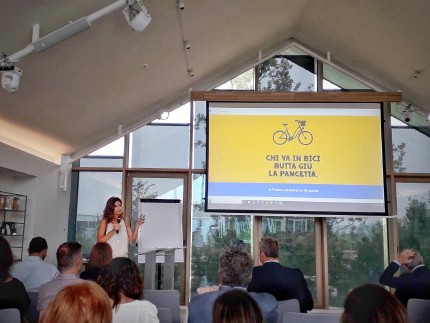 A woman giving a speech to a croud with a projection screen showing a bicycle and an Italian slogan A woman giving a speech to a croud with a projection screen showing a bicycle and an Italian slogan