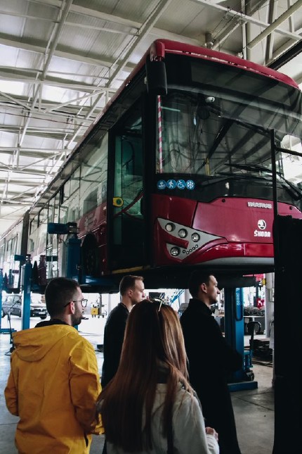 People looking at a trolleybus that is being suspended by a jack 