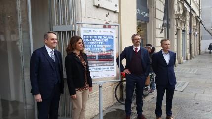 Four government officials standing in front of poster announcing national trolleybus day in Parma. Woman trying to unlock her bicycle directly behind them. Four government officials standing in front of poster announcing national trolleybus day in Parma. Woman trying to unlock her bicycle directly behind them.